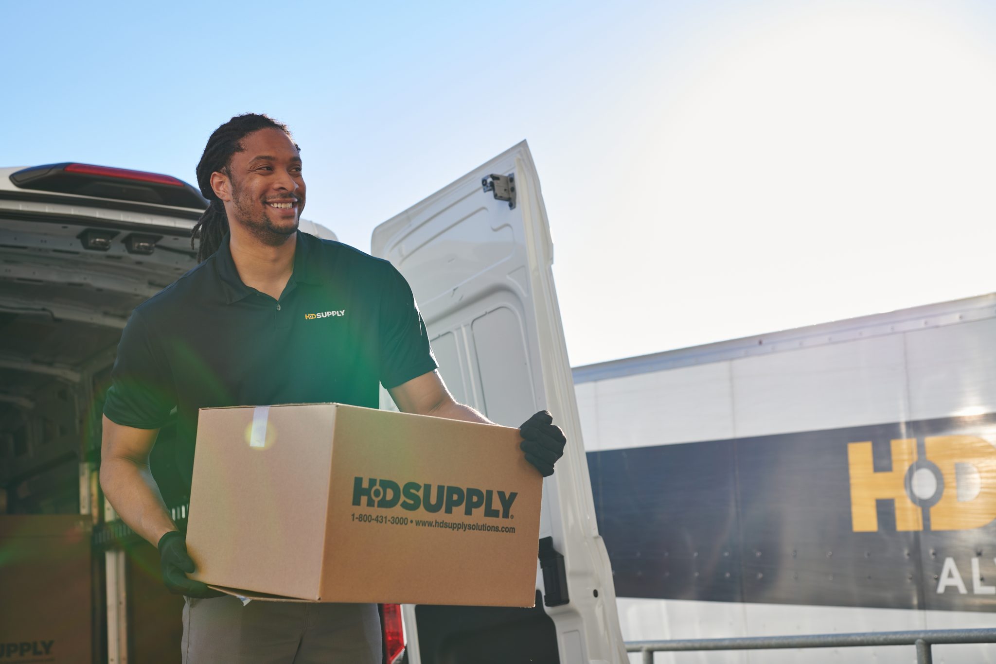 HD Supply delivery driver carrying HD Supply-branded box with HD Supply-branded deliver truck in background with back door lifted, featuring a sunrise sky